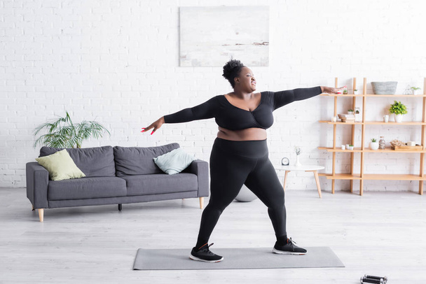 volledige lengte van tevreden Afrikaanse amerikaanse overgewicht vrouw in sportkleding oefening thuis - Foto, afbeelding
