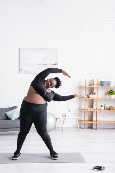 longitud completa de la alegre mujer afroamericana con sobrepeso en ropa deportiva haciendo ejercicio en casa - Foto, Imagen