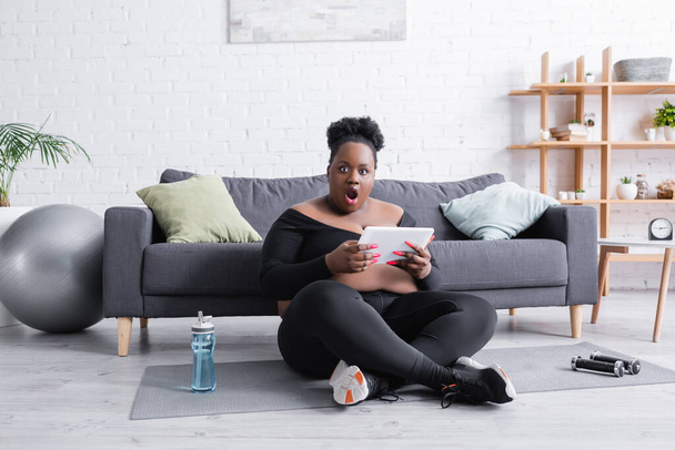 sorprendido afroamericano más tamaño mujer sosteniendo tableta digital en sala de estar - Foto, imagen