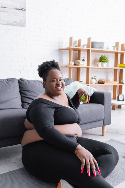 joyful african american plus size woman in sportswear holding green apple  - Photo, Image