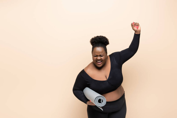 asombrado afroamericano más tamaño mujer en ropa deportiva celebración de la estera de fitness aislado en beige  - Foto, imagen