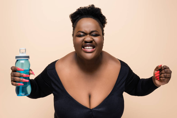 szczęśliwy african amerykański plus rozmiar kobieta w odzież sportowa gospodarstwa sportowe butelka odizolowany na beżowy  - Zdjęcie, obraz