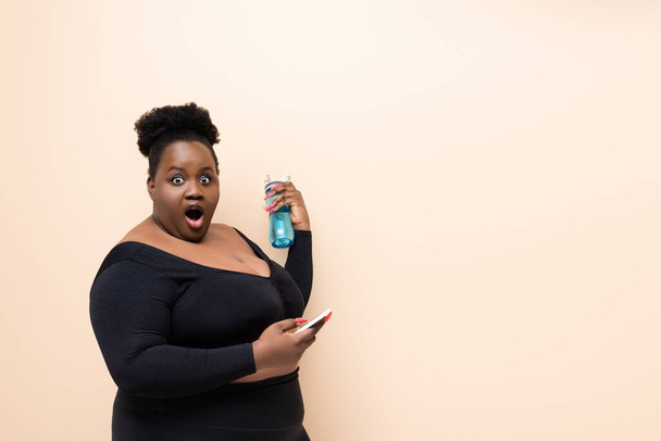 wstrząśnięty Afryki plus rozmiar kobieta w odzieży sportowej trzyma sportowe butelki i smartfona izolowane na beżu  - Zdjęcie, obraz
