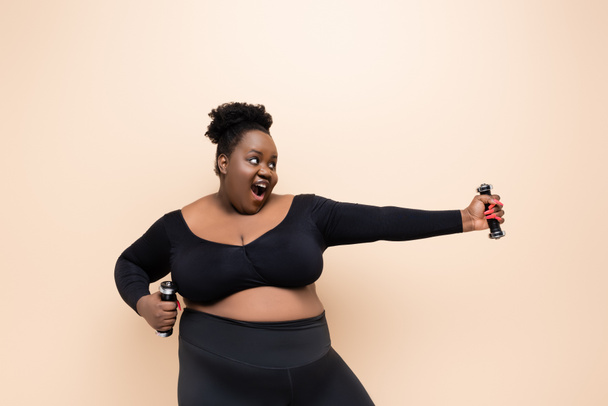 excited african american plus size woman in sportswear holding dumbbells isolated on beige  - Photo, Image