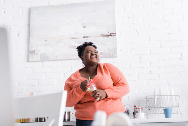 陽気なアフリカ系アメリカ人プラスサイズの女性保持プラスチックカップでおいしいスムージーでキッチン  - 写真・画像