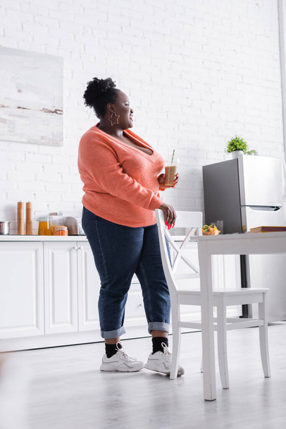 ευχαριστημένη αφρικανή αμερικανίδα plus size γυναίκα κρατώντας πλαστικό κύπελλο με νόστιμο smoothie στην κουζίνα  - Φωτογραφία, εικόνα