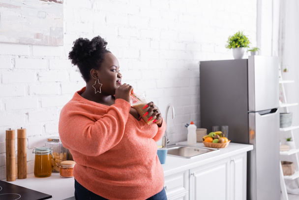 complacido afroamericano más tamaño mujer sosteniendo taza de plástico y bebiendo sabroso batido en la cocina  - Foto, Imagen