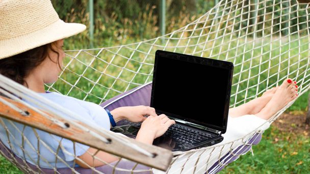 Back view young woman in summer hat student typing using laptop for e learning distance training course, lying on hammock outdoors. Working online at home. Over shoulder close up mock up screen view - Photo, Image