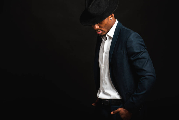 Πορτρέτο των ελκυστικών, χαριτωμένο, κομψό επαγγελματικό Αφροαμερικανός επιχειρηματίας σε κομψό κοστούμι, λευκό πουκάμισο και μαύρο καπέλο, απομονώνονται σε σκούρο φόντο. Χαμηλό κλειδί. Επιλεκτική εστίαση - Φωτογραφία, εικόνα