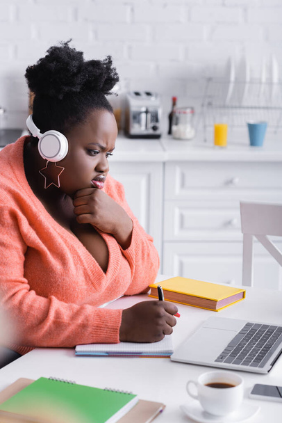 afro-américaine plus femme taille écoute podcast dans les écouteurs et en utilisant un ordinateur portable - Photo, image