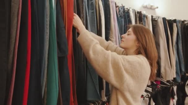Krásná mladá dívka nakupování a třídění přes oblečení na stojanu ve velkém obchodě - Záběry, video