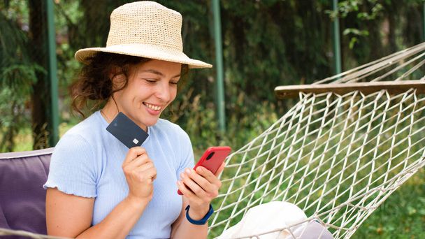 Szczęśliwa młoda kobieta w słomkowym kapeluszu z kartą kredytową banku, przy użyciu smartfona, dokonywania płatności internetowych, zakupy online, rezerwacja hotelu, bilety, zamówić taksówkę za pośrednictwem aplikacji, relaks w hamaku. Bankowość internetowa - Zdjęcie, obraz