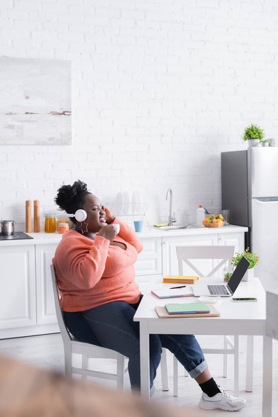 ευτυχισμένη Αφροαμερικανός συν μέγεθος γυναίκα στα ακουστικά κρατώντας κύπελλο στην κουζίνα  - Φωτογραφία, εικόνα