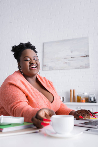 feliz afro-americano plus size mulher atingindo xícara de café em primeiro plano borrado  - Foto, Imagem