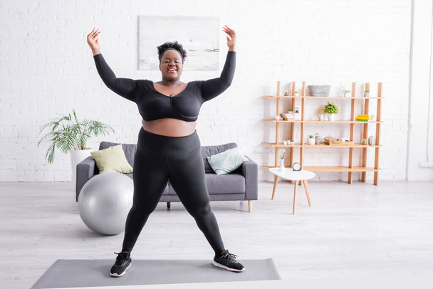 volledige lengte van vrolijke Afrikaans-Amerikaanse plus size vrouw in sportkleding training op fitness mat  - Foto, afbeelding