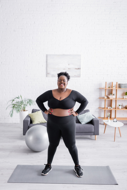 longitud completa de afroamericano más mujer de tamaño en ropa deportiva de pie con las manos en las caderas en la estera de fitness  - Foto, imagen