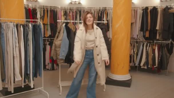 Elegante chica joven en pantalones vaqueros hipster está de compras en una tienda de ropa y está girando alrededor de probar un nuevo abrigo - Metraje, vídeo