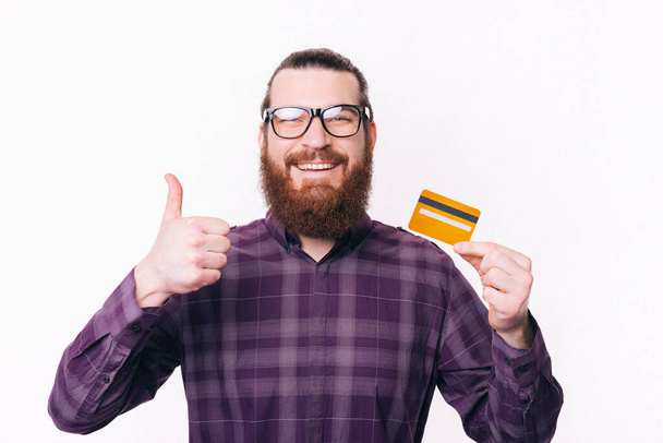 Portrait d'un bel homme portant des lunettes de vue tenant la carte de crédit et montrant pouce vers le haut. - Photo, image