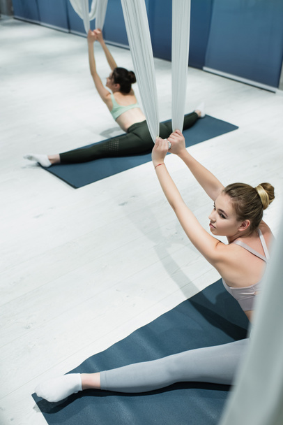 wysoki kąt widzenia młodych kobiet rozciągających się paskami do jogi muchowej podczas siedzenia na matach fitness - Zdjęcie, obraz