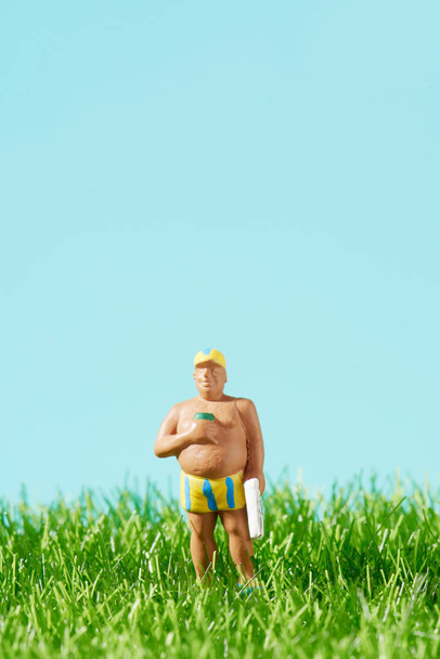 Крупный план миниатюрного человека в полосатом купальнике, держащего полотенце в одной руке и стакан в другой руке, стоящего на траве, на синем фоне с небольшим пустым местом наверху - Фото, изображение