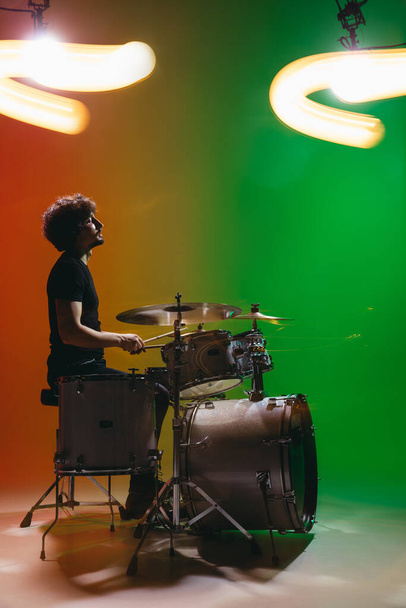 Porträt eines jungen Schlagzeugers, der isoliert auf grün-rotem Hintergrund in Neon spielt - Foto, Bild