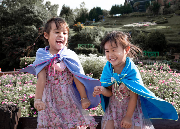Dwie małe dziewczynki w szal gra superbohatera uśmiech szczęśliwie w ogrodzie. - Zdjęcie, obraz
