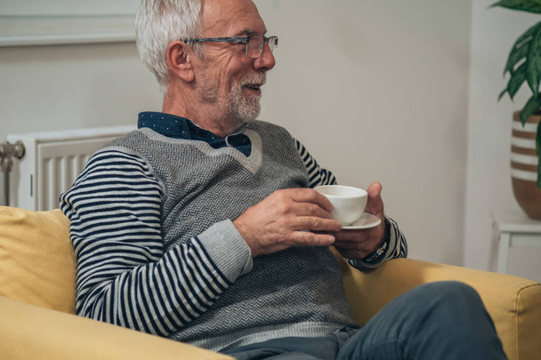 Hombre mayor guapo hablando y sosteniendo una taza de té en casa. Retrato de un hombre mayor con cabello gris y barba sentado en un sillón amarillo y bebiendo una taza de café mientras discute con alguien en casa - Foto, Imagen