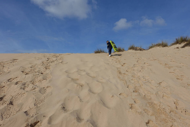 Menino levando sua prancha de boogie para o topo das dunas de areia para deslizar para baixo - Foto, Imagem