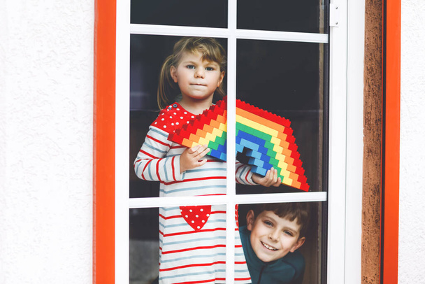 Toddler lány és kisfiú ablakról ablakra létre szivárvány színes műanyag blokkok alatt pandémiás koronavírus karantén. Két gyermek, testvérek létre, és festeni szivárványok szerte a világon, mint a jel. - Fotó, kép