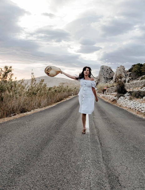 A young Croatian woman in a white dress enjoying the wind on the road through a valley during sunset - Φωτογραφία, εικόνα