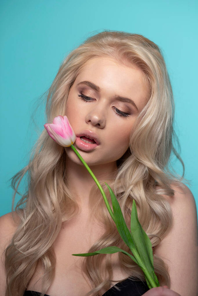 Sinnliche blonde junge Frau mit geschlossenen Augen, die seine Lippen mit rosa Tulpe vor blauem Hintergrund berührt. Studioporträt. - Foto, Bild