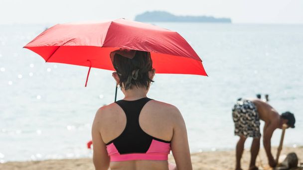 Egy női turista egy piros esernyőt tart, hogy megvédje magát a nap melegétől, míg a strandon van.. - Fotó, kép