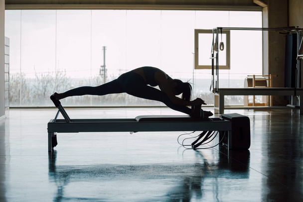 Fit εκπαιδεύτρια εξάσκηση Pilates ασκήσεις ρουτίνας σε ένα Reformer Bed Machine, λεπτή γυναίκα που εργάζονται έξω τεντώνοντας τα πόδια, κάνει ένα split, σε ένα μεγάλο γυμναστήριο Fitness με μεγάλα παράθυρα και θέα στο δρόμο. - Φωτογραφία, εικόνα