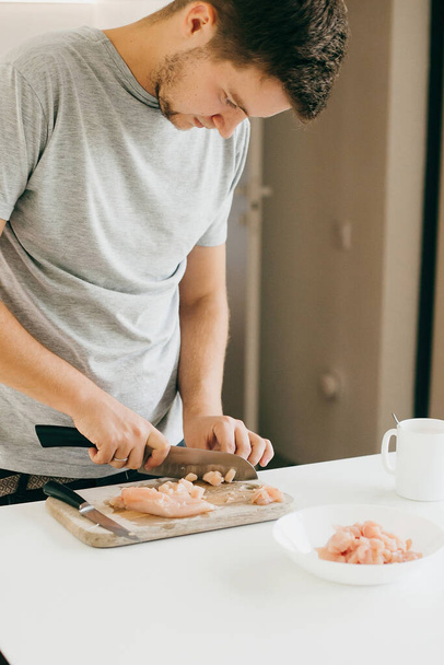 Young man cutting chicken fillet with knife on wooden board in modern white kitchen. Process of cooking and preparing meat, hands close up. Home cooking concept - Photo, Image