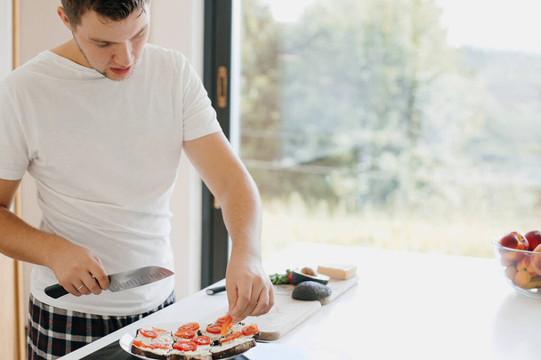 Nuori mies laittaa tomaatti viipaleita leipää ja tehdä avokado paahtoleipää moderni valkoinen keittiö. Maljapuhe. Terveellinen syöminen ja kotiruoanlaitto käsite - Valokuva, kuva