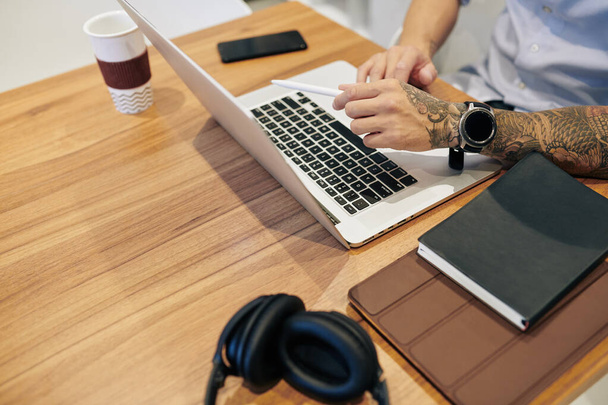 Крупный план бизнесмена, работающего на ноутбуке за столом с учебниками, наушниками, смартфоном и чашкой кофе - Фото, изображение
