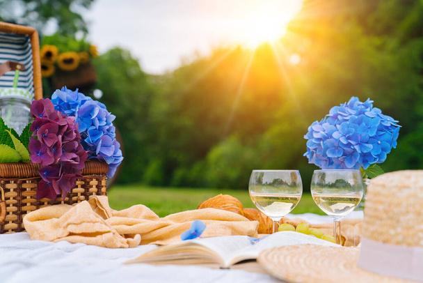 Nyári piknik napsütéses napon kenyér, gyümölcs, csokor hortenzia virágok, szemüveg bor, szalma kalap, könyv és ukulele. Piknik kosár a fűben, étellel és itallal, fehér kötött takarón. Szelektív fókusz. - Fotó, kép