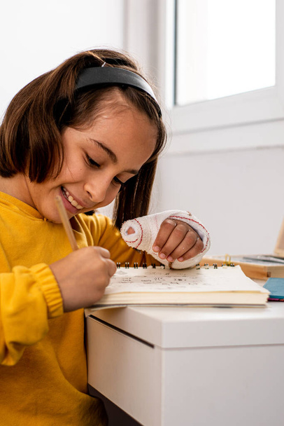 Petite fille caucasienne, avec un plâtre dans une main, portant un sweat-shirt jaune, faisant des croquis à son bureau très heureux et souriant, avec des matériaux de dessin, un carnet de croquis et une tablette. - Photo, image