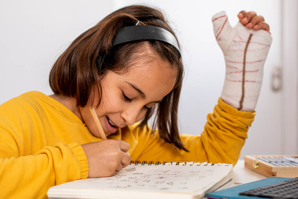 Petite fille caucasienne, avec un plâtre dans une main, portant un sweat-shirt jaune, faisant des croquis à son bureau très heureux et souriant, avec des matériaux de dessin, un carnet de croquis et une tablette. - Photo, image