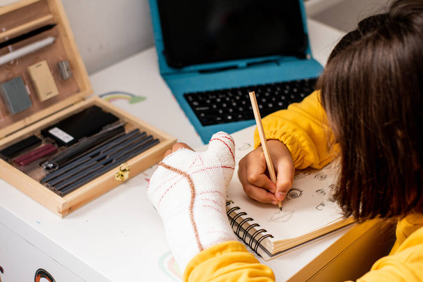 Detalhe de uma garota com um gesso em uma mão, vestindo um pulôver amarelo, desenhando esboços em seu caderno de esboços, em sua mesa de quarto, com materiais de desenho e um tablet. - Foto, Imagem