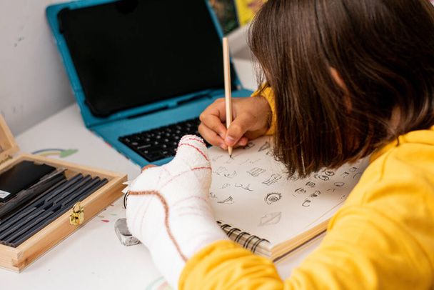 Detailfoto van een meisje met een gips in één hand, met een gele trui aan, schetsen in haar schetsboek, op haar slaapkamerbureau, met tekenmateriaal en een tablet. - Foto, afbeelding
