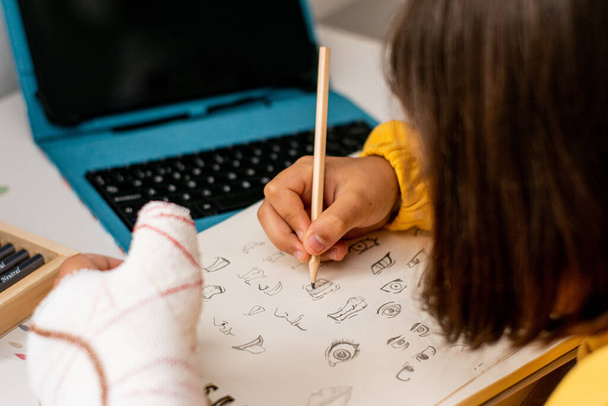 Detailfoto van een meisje met een gips in één hand, met een gele trui aan, schetsen in haar schetsboek, op haar slaapkamerbureau, met tekenmateriaal en een tablet. - Foto, afbeelding