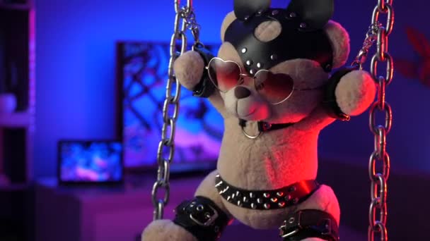 Teddy bear suspended from chains in bdsm accessories in neon light - Footage, Video