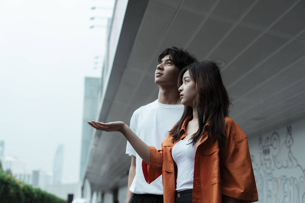 Nastoletnia para stojąca przy markizie centrum handlowego, dziewczyna wypuszczająca rękę, by sprawdzić deszcz, oboje patrzą na niebo.. - Zdjęcie, obraz