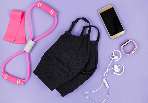 Спортивная одежда и аксессуары для женщин на фиолетовом фоне. Спортивная мода с футболкой, резинками, наушниками, телефоном, бутылкой. Концепция здорового и активного образа жизни - Фото, изображение