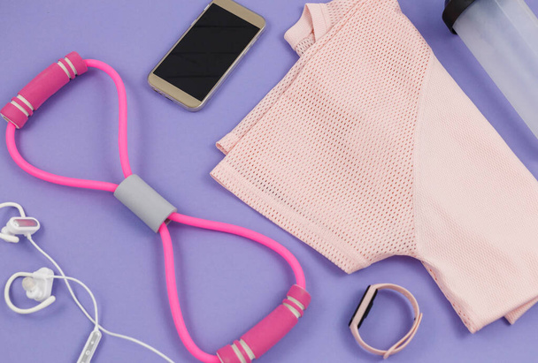 Vêtements et accessoires de fitness pour femmes sur fond violet. Mode sportive avec un t-shirt, bandes élastiques, écouteurs, téléphone, bouteille. Mode de vie sain et actif concept - Photo, image