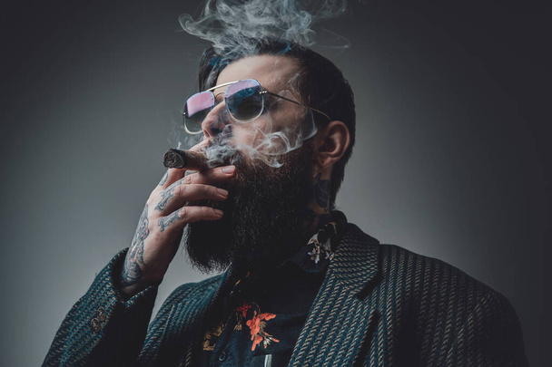 Cool en zelfverzekerde man rookt sigaar in grijze achtergrond - Foto, afbeelding
