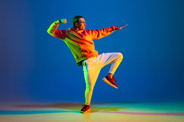 Young man dancing hip-hop in stylish attire on blue background in neon light - Photo, Image