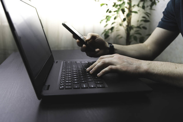 Adam ofiste ya da evde akıllı telefon ve dizüstü bilgisayar kullanıyor. Erkeklerin klavye yazarken ve cep telefonu kullanırken yan görüntü görüntüsü - Fotoğraf, Görsel