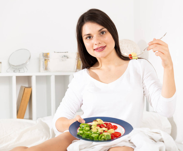 Csinos nő fehér ruhában eszik zöldséges salátát az ágyban - Fotó, kép